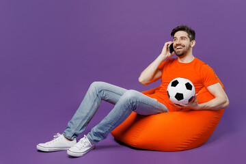 Full size young fan man he wear orange t-shirt cheer up support football team hold soccer ball watch tv live stream sit in bag chair talk speak on mobile cell phone isolated on plain purple background