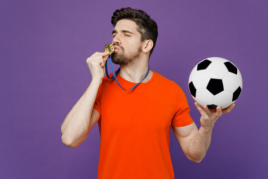 Young exultant jubilant happy fan man he wears orange t-shirt cheer up support football sport team hold in hand soccer ball kiss medal on neck watch tv live stream isolated on plain purple background.
