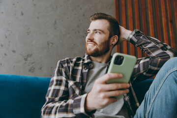 Top view young man 20s he wears brown shirt hold in hand use mobile cell phone look aside sitting on blue sofa in own living room apartment stay home indoor flat on weekends. People lifestyle concept.