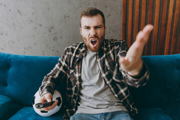 Fototapeta Young mad angry man fan in brown shirt hold remote controller hold head cheer up support football team sit on blue sofa with soccer ball rest watch tv indoors room gray wall. Sport leisure concept. obraz