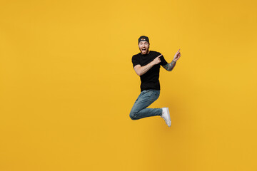 Fototapeta na wymiar Full body young bearded tattooed man 20s he wears casual black t-shirt cap jump high indicate point index finger aside on workspace area isolated on plain yellow wall background. Tattoo translate fun.