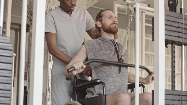 Tilt up of hard working young Caucasian man in wheelchair doing lat pulldown exercises in gym during personal training with African American physiotherapy coach