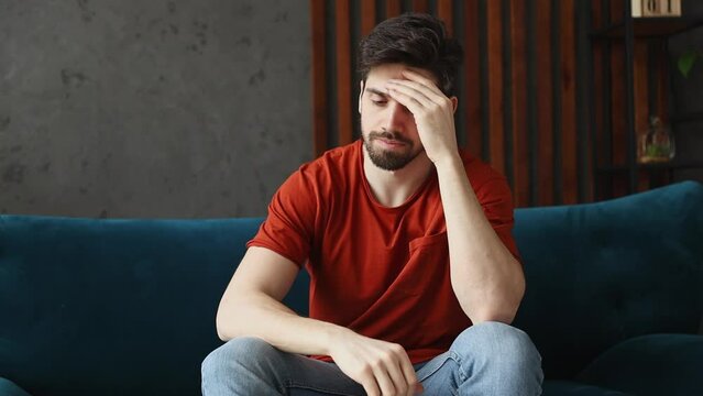 Young sad puzzled pensive gloomy man wears red t-shirt put hand on head prop up on chin iterates over solution options sits on blue sofa stay at home flat spend free time in room indoors grey wall