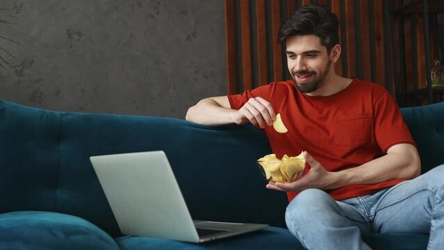 Young smiling man wears red t-shirt watch movie film on laptop pc computer eat chips sits on blue sofa stay at home rest relax spend free time in living room indoors grey wall. People lounge concept