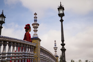 Fototapeta na wymiar Beautiful teenage woman dancing flamenco on a bridge overlooking a famous square in Seville She is wearing a red dress with a ruffle and red flowers on her head. Flamenco cultural heritage of humanity