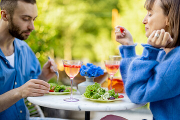 Young and cheerful couple have fun, talking and eating healthy salad while sitting together at backyard during summer time