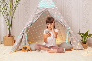 Portrait of little girl sitting at home in teepee and playing on the phone, holding cell phone with...