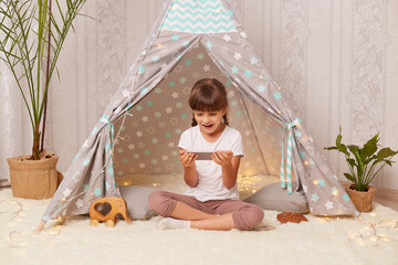 Indoor shot of shocked little girl with pigtails wearing white t shirt posing in wigwam at home and using phone, looking at display with open mouth, sees something astonishing in Internet. © sementsova321