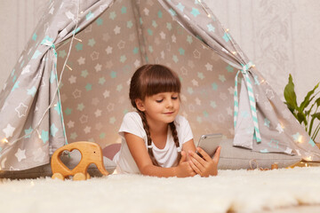 Satisfied delighted little girl with pigtails wearing white t shirt posing in wigwam at home and using phone, watching cartoons or having video call, expressing positive emotions. © sementsova321
