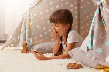 Profile portrait of sleepy tired little girl with pigtails wearing white t shirt posing in wigwam at home and using phone, checking social networks, being sleepless, yawning, covering mouth with palm. © sementsova321