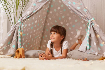 horizontal shot of dreamy little girl with pigtails wearing white t shirt posing in wigwam at home, kid holding mobile phone, looking away with positive smile, enjoying leisure time in peetee tent.