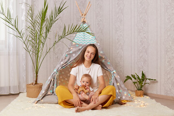 Horizontal shot of smiling beautiful woman sitting with her toddler daughter, mother playing at home in wigwam with her kid while sitting on the floor on soft carpet. © sementsova321