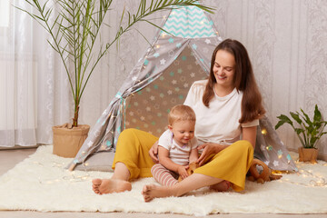 Portrait of mother playing at home in wigwam with her baby daughter while sitting on the floor, female looking at toddler kid with love and gentle, happy motherhood and childhood. © sementsova321
