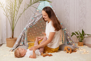 Indoor shot of young mother playing with with baby daughter on floor near kids wigwam, expressing positive emotions, spending time with cute kid in peetee tent at home. © sementsova321