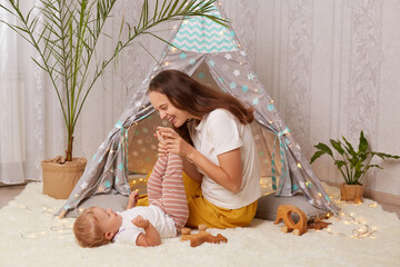 Indoor shot of smiling happy excited mother playing at home in peetee tent while sitting on the floor and having fun with infant toddler baby, mommy holding child's feet. © sementsova321