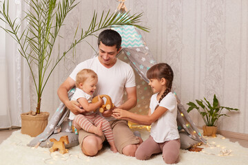 Indoor shot of happy father young man and his little daughters having fun and playing in wigwam in children room at home, play with toys, spending joyful time together. © sementsova321
