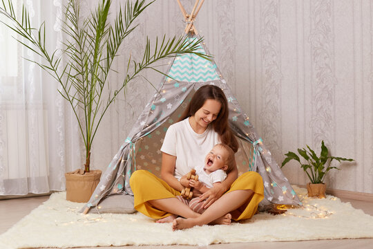 Image of happy laughing baby girl and her mother playing at home in wigwam while sitting on the floor, kid holding wooden toy, mommy enjoying her time with charming daughter.