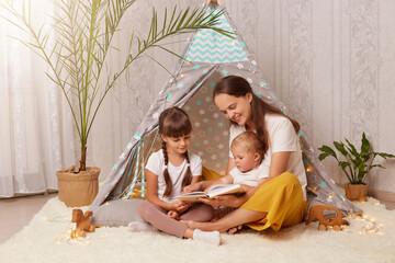 Caring mother and her daughters sitting near wigwam with book in hands, woman and kids playing at home, reading fairy tales and interesting stories, spending time together. © sementsova321