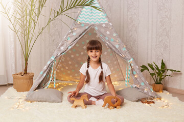 Image of cute happy little girl wearing white t shirt playing with wooden eco toys in teepee, charming female child with dark hair and braids enjoying while play in her new wigwam. © sementsova321