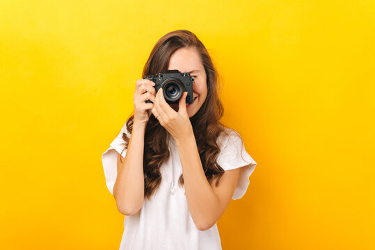 A young woman happily taking a photo with a black camera near a yellow background .