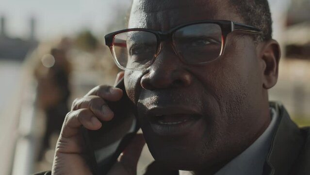 Close up shot of African American businessman in glasses talking on mobile phone while standing outdoors in city