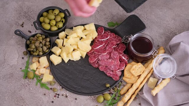 making meat and cheese antipasto plater - woman putting pieces of parmesan hard cheese on stone serving board at domestic kitchen