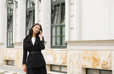 Fototapeta premium Successful businesswoman in a suit with a document goes to a meeting near a modern office building and talks on a smartphone