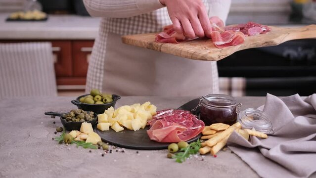 making meat and cheese antipasto plater - woman putting pieces of prosciutto ham on stone serving board at domestic kitchen