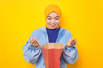 Excited young Asian woman in jeans jacket looking inside shopping bags isolated over yellow background