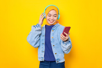 Dancing young Asian woman in jeans jacket wearing headphones for listening to music via mobile phone isolated over yellow background