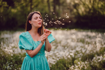 Smiling woman beautiful blowing on ripened dandelion in park. Girl in retro turquoise dress enjoying summer in countryside. Wishing, joy concept. Springtime, aesthetic portrait