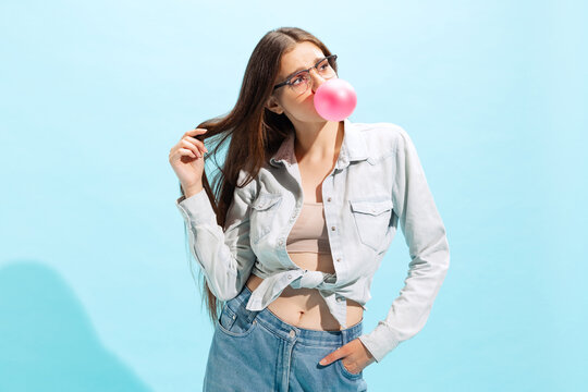 Portrait of young girl in casual clothes posing with pink bubblegum isolated over blue studio background.