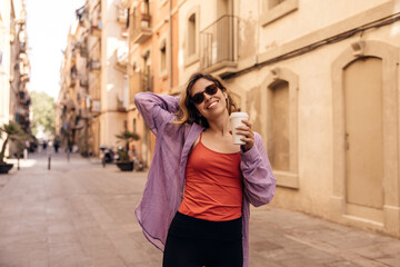 Happy young caucasian woman enjoys walking around city sunny weather. Brown-haired girl wears casual clothes and coffee in her hands. Summer playful mood concept