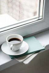 Coffee cup and books on windowsill. Cozy minimal still life against wet window. Rest and reading at rainy day.