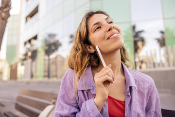 Enthusiastic european young lady looking away, holding stylus for tablet sitting outdoors....