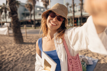 Smiling young caucasian woman taking selfie spending leisure time on beach in sunny weather. Blonde wears hat, sunglasses and shirt. Summer vacation concept