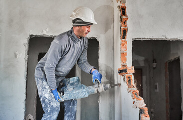 Side view male professional of demolition walls in construction helmet using perforator in mode of jackhammer and destroying interroom brick septum. Redevelopment, reconstruction of housing.