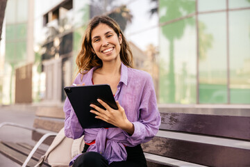 Positive young caucasian woman holding tablet smiling looking at camera outside. Brown-haired wears purple shirt to street. Social media addiction concept