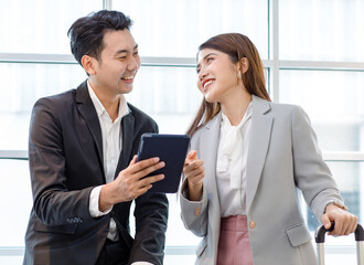 Asian young professional successful businessman and businesswoman colleague in formal suit standing together with trolley baggage in airport hallway waiting checking for business trip boarding time