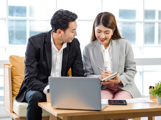 Millennial Asian young professional successful businessman employee in formal suit sitting on sofa smiling typing laptop computer while businesswoman colleague holding coffee cup helping advising