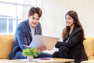 Millennial Asian professional successful male businessman and female businesswoman in formal suit sitting on sofa smiling discussing together while working with laptop notebook computer in office