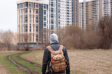 A young man with a hunting backpack is walking near a high building on the cold autumn weather holding a long rod. Back view