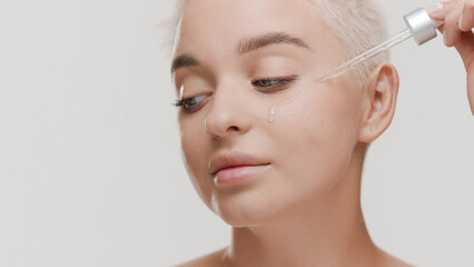 Fototapeta premium Attractive young slim blonde Caucasian woman drips clear serum from pipette on her cheek looking away on white background | Skincare serum commercial