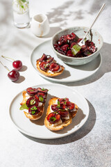 Toasts with soft cheese and baked with herbs sweet cherry berries on white concrete background. Roasted cherries. Delicious breakfast idea.