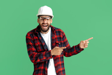 Happy bearded engineer or constructor man in casual outfit pointing finger away over green background.