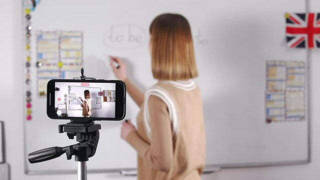 Competent female english teacher writing grammar rules on whiteboard while recording video on modern smartphone. Focus on cell phone screen. Blur background.