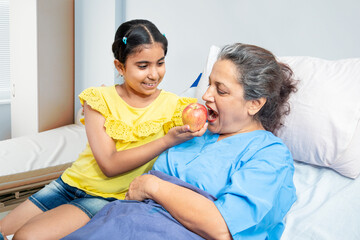 Little granddaughter help her grandmother patient eating apple in hospital bed. 