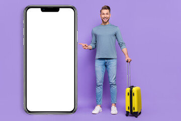 Photo of positive excited guy wear grey shirt holding suitcase pointing modern device empty space isolated violet color background