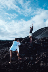 Young woman in a white shirt taking a photo of a young man on a rock in a volcanic landscape