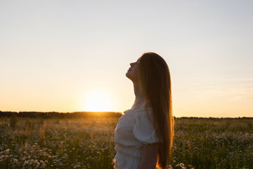 A beautiful redheaded girl is enjoying life in a field of daisies at sunset. A young woman with a wreath of daisies on her head smiles happily in nature.